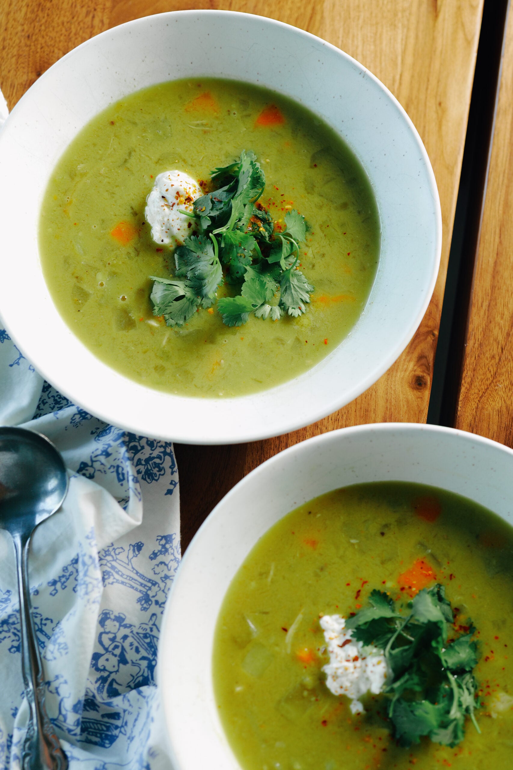 Two bowls of green vegetable soup topped with a dollop of cream, fresh cilantro, and red pepper flakes.