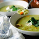 A spoonful of green vegetable soup held over a bowl topped with a dollop of cream and fresh herbs.
