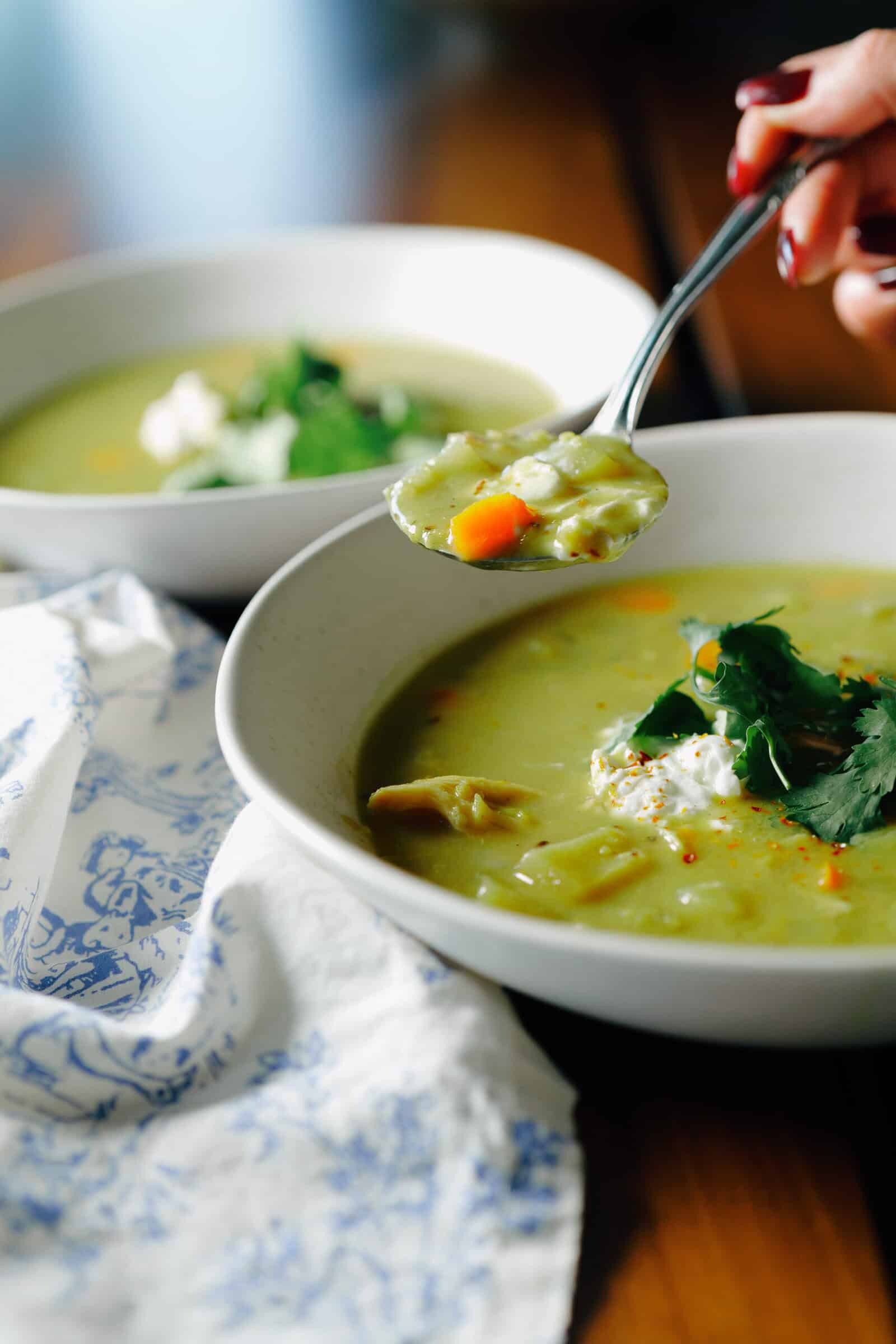 A spoonful of green vegetable soup held over a bowl topped with a dollop of cream and fresh herbs.